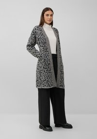 Femme portant un cardigan ouvert à imprimé léopard gris, un pull col roulé blanc, un pantalon large noir et des mocassins noirs se tient sur un fond gris.