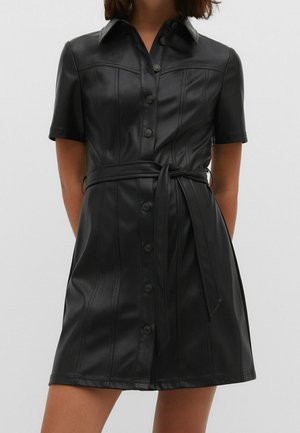 Zwarte faux leren korte mouwen hemdjurk met knopen aan de voorkant en een gestrikte tailleband, gedragen door een persoon tegen een effen achtergrond.