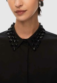 Camicia nera con colletto a punta, decorata con gemme nere lungo il bordo del colletto, caratterizzata da una texture liscia e un design strutturato.