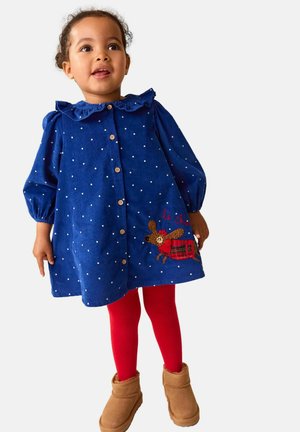 Blauwe corduroy jurk met witte stippen, ruches aan de kraag, knopen aan de voorkant en geborduurde rendieren. Gecombineerd met rode maillots en een paar bruine laarzen.