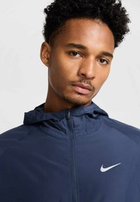 Granatowa kurtka zapinana na zamek, wykonana z lekkiego materiału, z kapturem i małym białym logo Nike po lewej stronie. Gładka tekstura, nowoczesny krój.