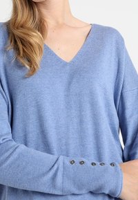 Jasnoniebieski sweter w serek o luźnym kroju, wykonany z miękkiego materiału. Posiada guziki na mankietach rękawów. Gładka powierzchnia.