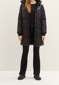 Kobieta w długiej czarnej kurtce puchowej, nosząca czarne prążkowane spodnie i czarne masywne buty, stojąca na jednolitym jasnym tle.