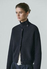 Frau trägt eine schwarze, gerippte, zum Knöpfen geeignete Strickjacke mit hohem Kragen und langen Ärmeln, blickt seitlich vor einem schlichten Hintergrund.