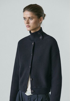 Kvinde iført en sort ribbet cardigan med knapper, høj krave og lange ærmer, kigger til siden mod en ensfarvet baggrund.