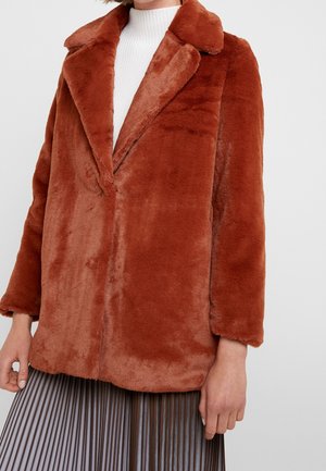 Donna che indossa una giacca in pelliccia sintetica color ruggine sopra un dolcevita bianco e una gonna scura plissettata, in piedi davanti a uno sfondo semplice.