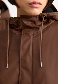 Hnedá, ľahká, vodeodolná bunda s vysokým golierom, sťahovacou šnúrkou a hladkým povrchom. Detaily zahŕňajú jemné stehy.