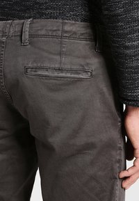 Pantalon en coton gris avec une poche arrière à ouverture horizontale, texture lisse et détails de couture minimaux.