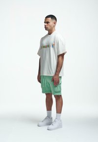 Cremfarbendes Oversized-T-Shirt mit buntem Text, kombiniert mit mintgrünen Cargo-Shorts und weißen Sneakern. Das Outfit bietet eine lässige Passform und weiche Materialien.