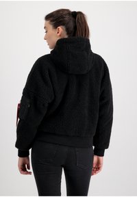 Svart, kort jacka med huva i texturerad fleece, med ribbad nederkant och mudd; har en exteriör ficka på vänster arm.