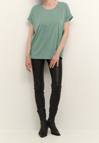 Tyrkysově zelené oversized tričko s ohrnutými rukávy, spárované s lesklými černými koženými kalhotami a nadkolenními špičatými botami na klínkovém podpatku.