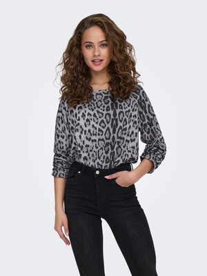 Femme aux cheveux bouclés portant une chemise à manches longues gris et noir imprimée léopard et un jean noir taille haute, debout avec une main dans la poche.