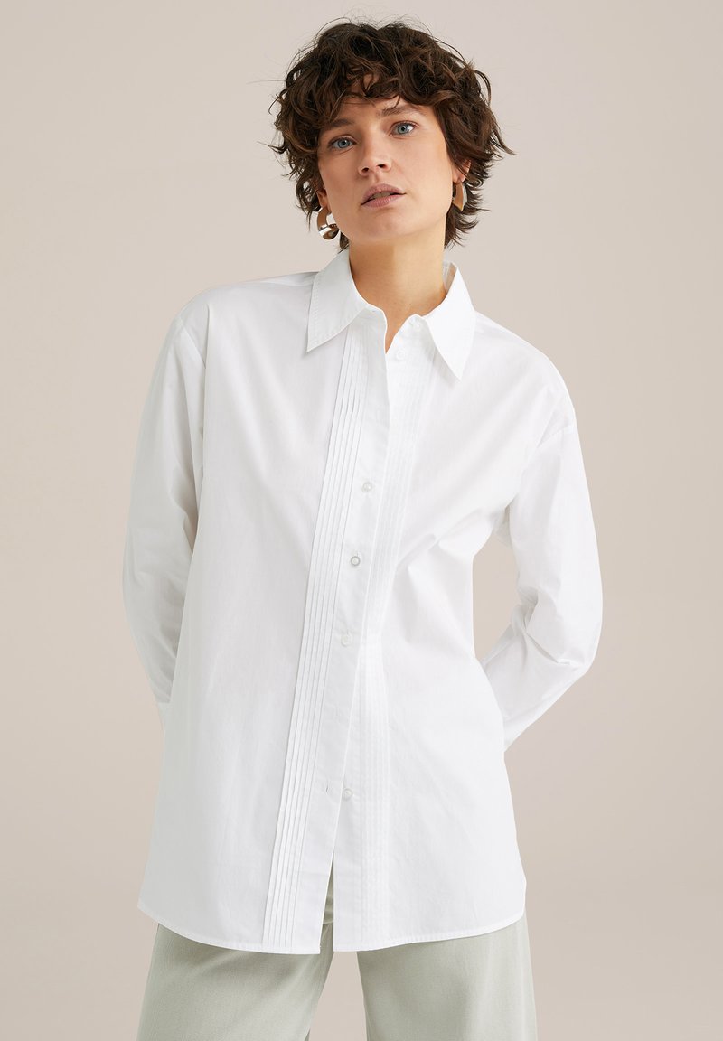 Biela košeľa s dlhým rukávom a klasickým golierom, s vertikálnymi prehnutiami na prednej strane a voľným strihom. Hladká bavlnená textura.
