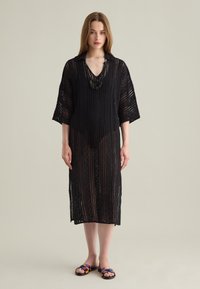 Eine Frau steht in einem schwarzen gehäkelten Midi-Kleid über einem schwarzen Bodysuit und bunten flachen Sandalen vor einem schlichten hellen Hintergrund.