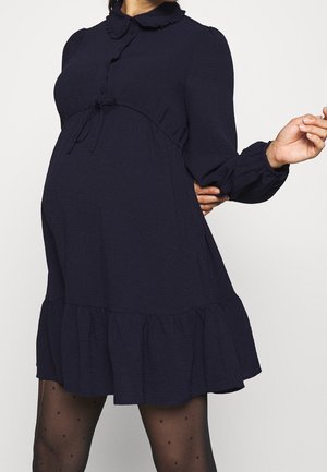 Femme enceinte portant une robe bleu marine à manches longues avec col et ourlet à volants, assortie à des collants noirs transparents à pois.