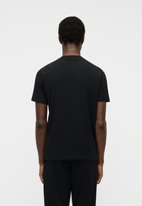 Άνδρας φαίνεται από πίσω φορόντας μια απλή μαύρη κοντομάνικη μπλούζα και μαύρο παντελόνι, με φόντο ένα λευκό τοίχο.