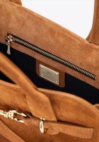 Sac en daim marron avec une poche zippée, quincaillerie dorée, un intérieur en tissu noir et une plaque de marque en métal à l'intérieur.