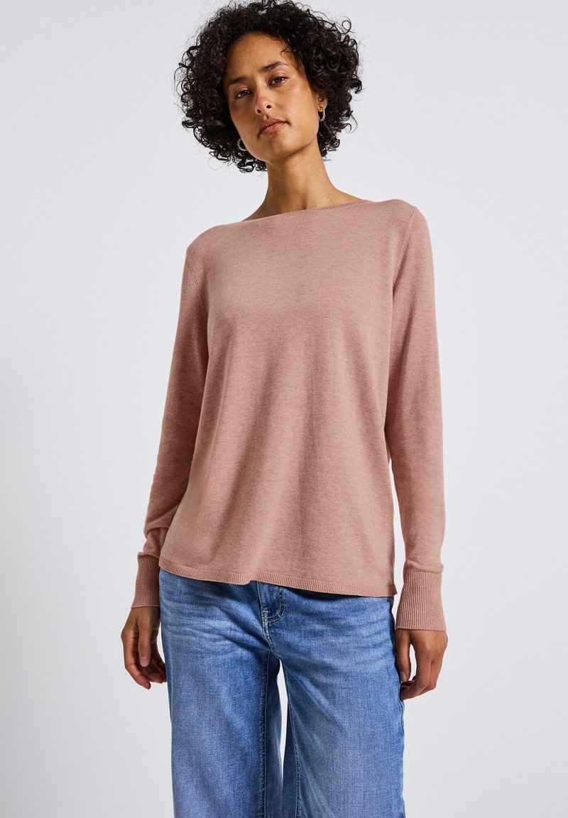 Helles rosa Sweater mit weitem Ausschnitt und langen Ärmeln, aus weichem Stoff gefertigt, mit gerippten Bündchen und lässiger Passform. Kombiniert mit blauen Jeans.
