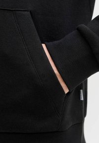 Sweat-shirt noir avec poche kangourou à l'avant, poignets et ourlet côtelés. Texture lisse avec des détails de couture visibles.