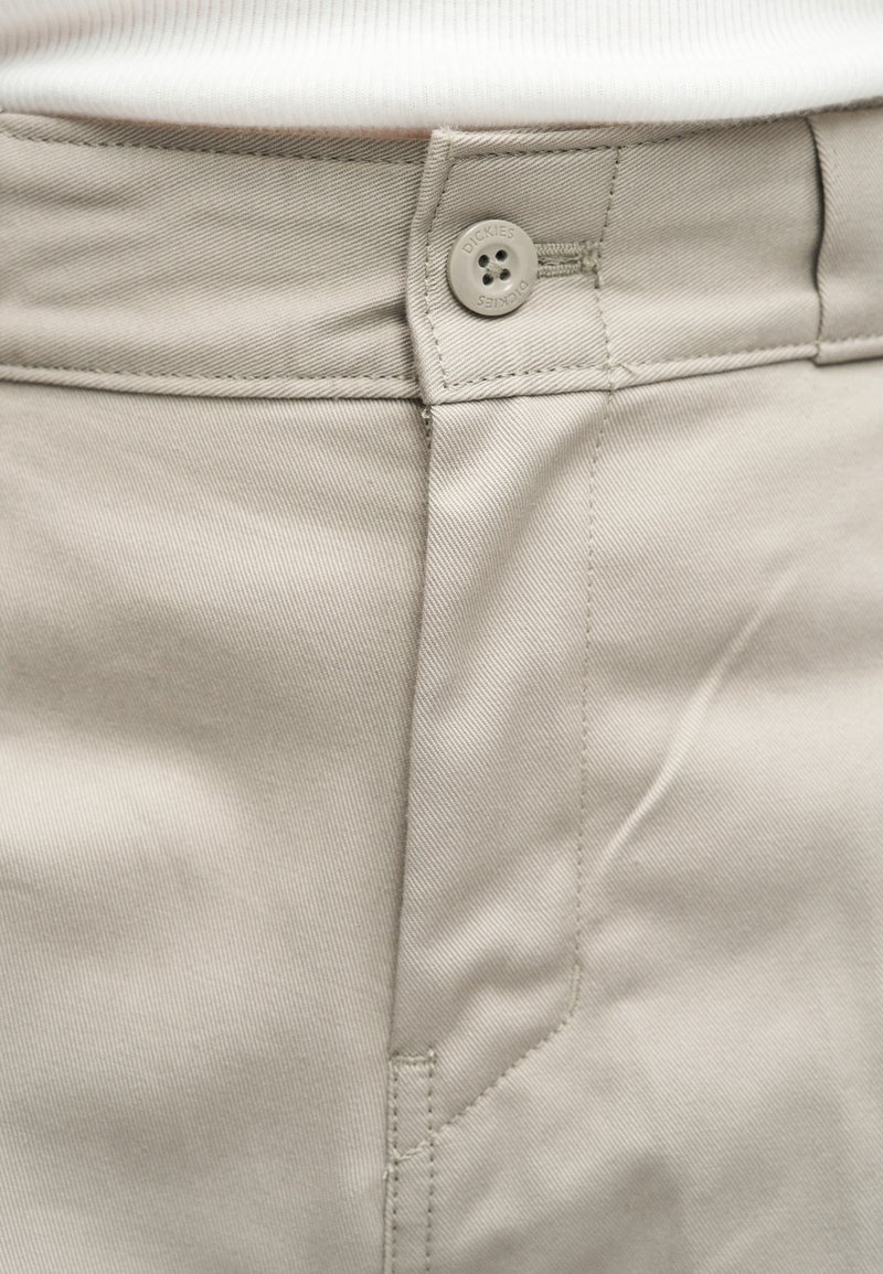 Gros plan sur la ceinture d'un pantalon beige avec un bouton marqué « Dickies » et des détails de couture visibles, porté avec une chemise blanche.