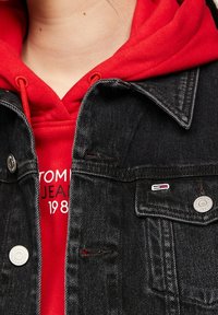 Personne portant un hoodie rouge sous une veste en denim noir avec des boutons argentés et un petit logo rectangulaire sur la poche de poitrine.