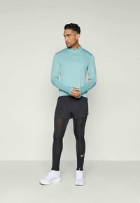 Langärmliges, mintgrünes Sportshirt mit einem Reißverschluss am Hals, kombiniert mit schwarzen Shorts und schwarzen Leggings. Weiße Laufschuhe mit Logo-Akzenten.