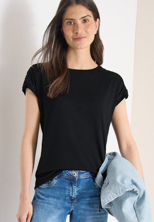 Jeune femme aux longs cheveux bruns portant un t-shirt noir et un jean bleu, tenant une veste en denim bleu clair sous son bras gauche.