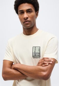 Νέος άνδρας με κοντά σγουρά μαλλιά που φοράει κρεμ μπλουζάκι με μικρό ορθογώνιο γραφικό και κείμενο "MOPD SPORTS CLUB", με τα χέρια σταυρωμένα.