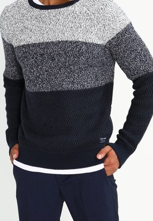 Mann trägt einen gestrickten Pullover mit horizontalen Farbblöcken in Hellgrau, Dunkelgrau und Marineblau, kombiniert mit dunkler Hose und weißem Unterhemd.