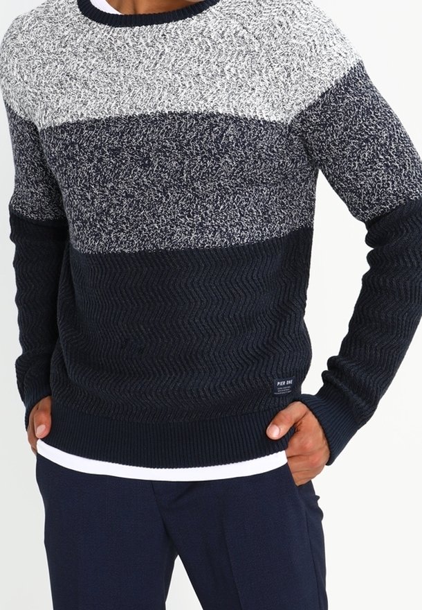 Homme portant un pull tricoté avec des blocs de couleurs horizontaux en gris clair, gris foncé et bleu marine, associé à un pantalon foncé et un sous-vêtement blanc.