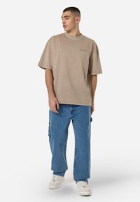 Beige, nagyméretű póló kerek nyakkivágással és rövid ujjakkal, világoskék farmernadrággal és fehér sportcipővel kombinálva, fehér háttér előtt állva.