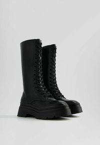Bottes montantes noires en simili cuir avec une semelle plateforme robuste, dotées d'un laçage prononcé et d'une finition lisse et brillante.