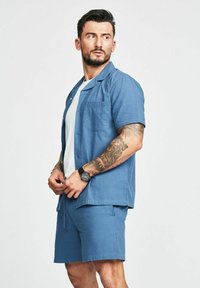 Camicia azzurra a maniche corte con texture abbinata a pantaloni corti coordinati. Presenta un colletto classico e una tasca a toppa, indossata con una t-shirt bianca sotto.