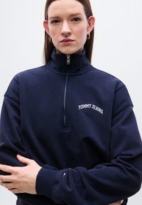 Ναυτικό μπλε φούτερ με φερμουάρ στο ένα τέταρτο, κατασκευασμένο από βαμβάκι, με μονόχρωμο σχέδιο, ριμπ μανσέτες και κεντημένο το "TOMMY JEANS" στο στήθος.