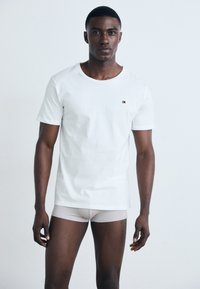 Hombre joven de pie, mirando hacia adelante, vistiendo una camiseta blanca lisa de cuello redondo y bóxers de color gris claro, contra un fondo blanco.