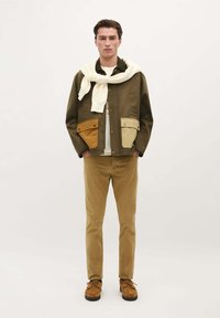 Veste bicolore verte et beige avec de grandes poches, pull blanc drapé autour du cou, pantalons en velours côtelé beige et chaussures marron.