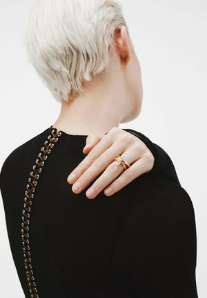 Persona con cabello corto rubio y camiseta negra, mostrando una mano con dos anillos de oro y un pequeño diamante, con la mano descansando sobre su hombro.