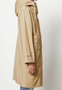 Person som bär en beige huvtröja med framfickor och knappar, visas från sidan mot en vit bakgrund.