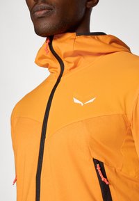 Πορτοκαλί μαλακή ζακέτα με μαύρους φερμουάρ και λεπτομέρειες. Διαθέτει ψηλό γιακά, υφασμάτινη επιφάνεια και μικρό άσπρο λογότυπο στο στήθος.