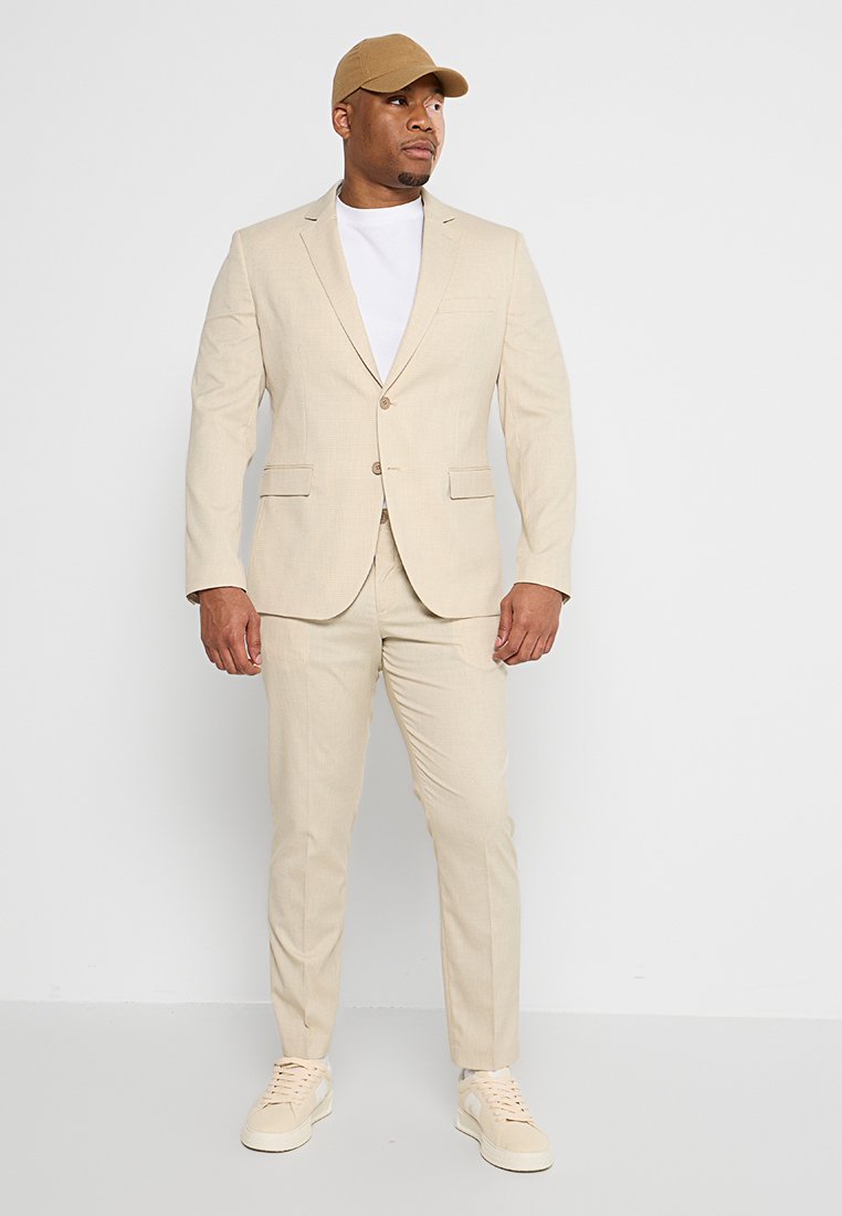 Beige Anzug mit strukturiertem Stoff, einreihiger Jacke, zwei vorderen Taschen und passender Hose, kombiniert mit einem weißen T-Shirt und beige Sneakern.