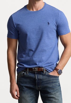 Niebieska koszulka z krótkim rękawem i okrągłym dekoltem, wykonana z teksturowanego materiału, z małym logo na lewej piersi. Noszona z dżinsami.