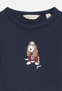 T-shirt bleu marine à col rond avec une impression d'un basset hound portant un pull rouge avec un logo "G" et du texte blanc en dessous.