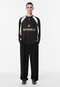 Schwarzes Langarmshirt mit weißen Akzenten und PlayStation 2-Logo, kombiniert mit schwarzen Hosen und schwarzen Sneakers.