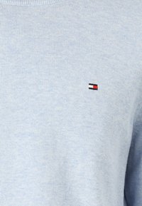 Svetlomodrá pletená látka s malým obdĺžnikovým logom s čiernymi, bielymi a červenými časťami vyšité na oblasti hrudníka.