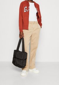 Vrouw in een roestrode zip-hoodie, witte top, beige broek en witte sneakers, met een zwart gewatteerd tote bag in één hand.