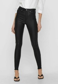 Leggings en simili cuir noir avec une taille haute, coupe slim et détails de couture visibles. Associés à une chemise blanche à boutons et des sandales.