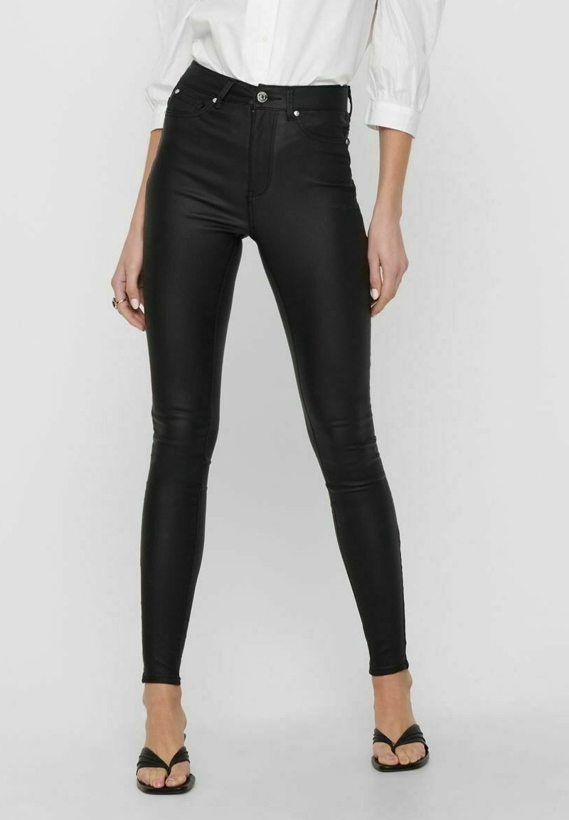 Leggings en simili cuir noir avec une taille haute, coupe slim et détails de couture visibles. Associés à une chemise blanche à boutons et des sandales.