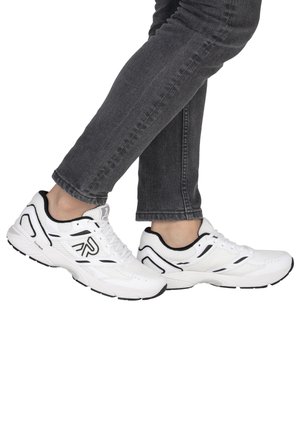 Person in schwarzen Jeans und weißen Sportschuhen mit schwarzen Akzenten, steht mit einem Fuß leicht hinter dem anderen erhöht.