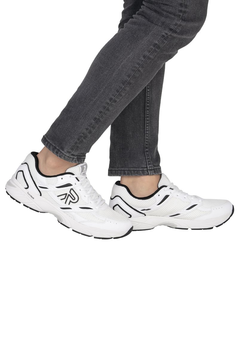 Personne portant un jean noir et des baskets blanches avec des accents noirs, se tenant avec un pied légèrement levé derrière l'autre.