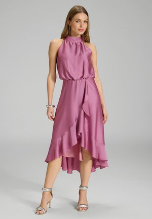 Halterneck pink satin dress with a fitted waist, flowing ruffled hem, and asymmetrical length; paired with silver heeled sandals.